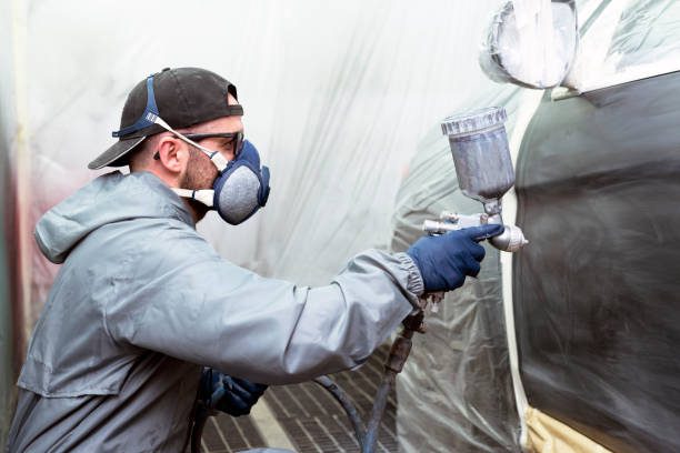 Car painter painting a car door with a mask on, gloves and work clothes